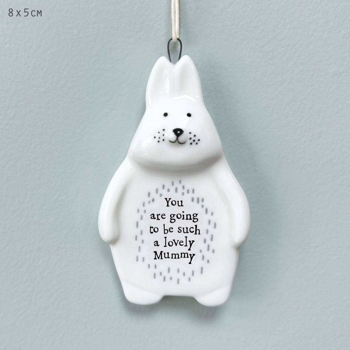 A white ceramic bunny ornament with a hanging loop at the top, featuring black facial details and the printed message 'You are going to be such a lovely Mummy' with small gray dotted patterns on its belly. The background is a soft light blue-gray wall, and '8 x 5cm' is printed in the upper left corner.