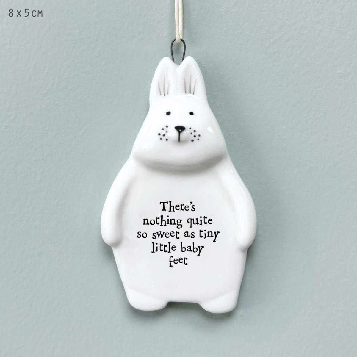 A white ceramic bunny ornament with black printed text reading 'There's nothing quite so sweet as tiny little baby feet' and a hanging loop at the top, shown against a pale blue-gray wall. Small text in the upper left says 8 x 5 cm.