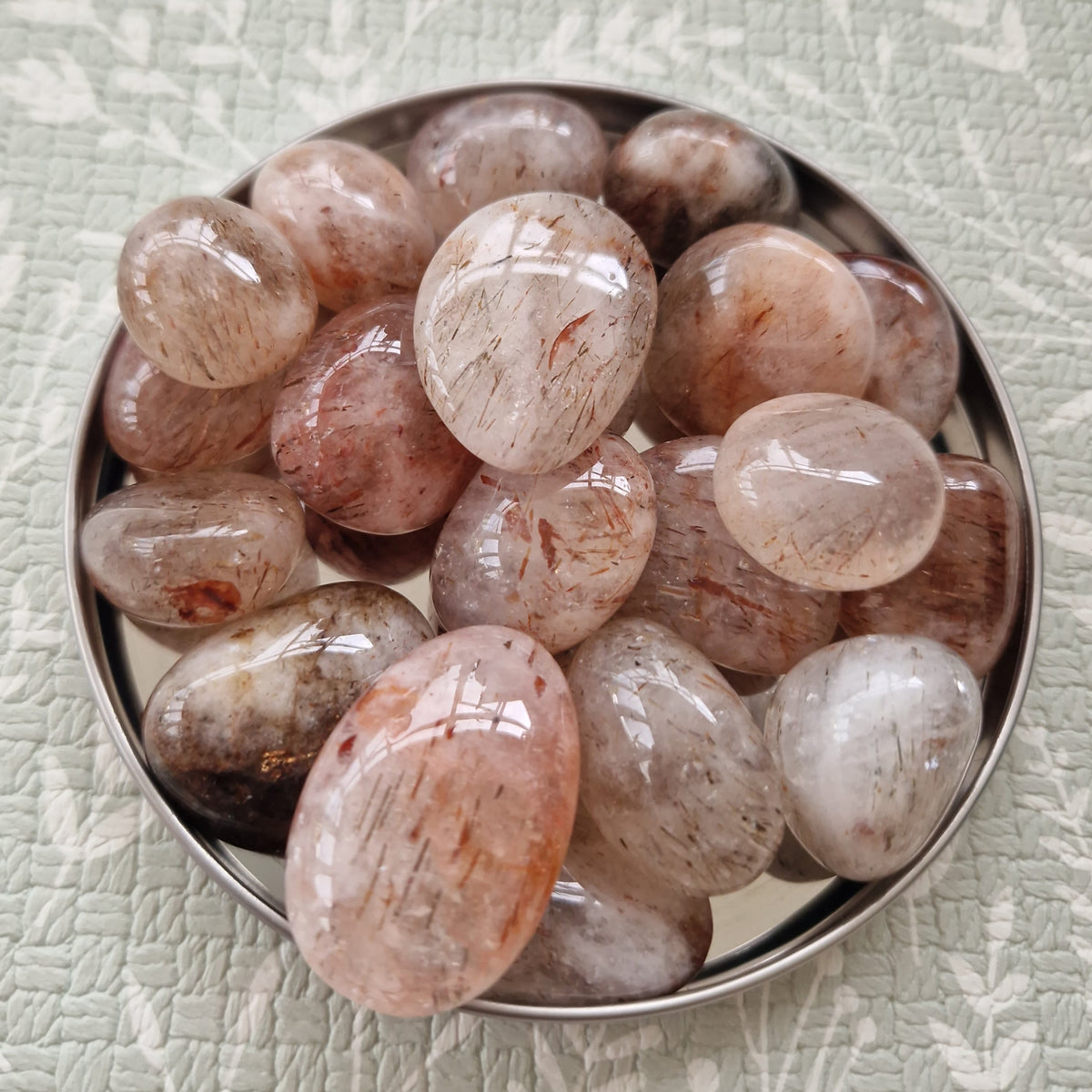 A pile of polished peach and white quartz crystal gemstones with fine inclusions, displayed in a small silver metal bowl on a textured sage green fabric background.