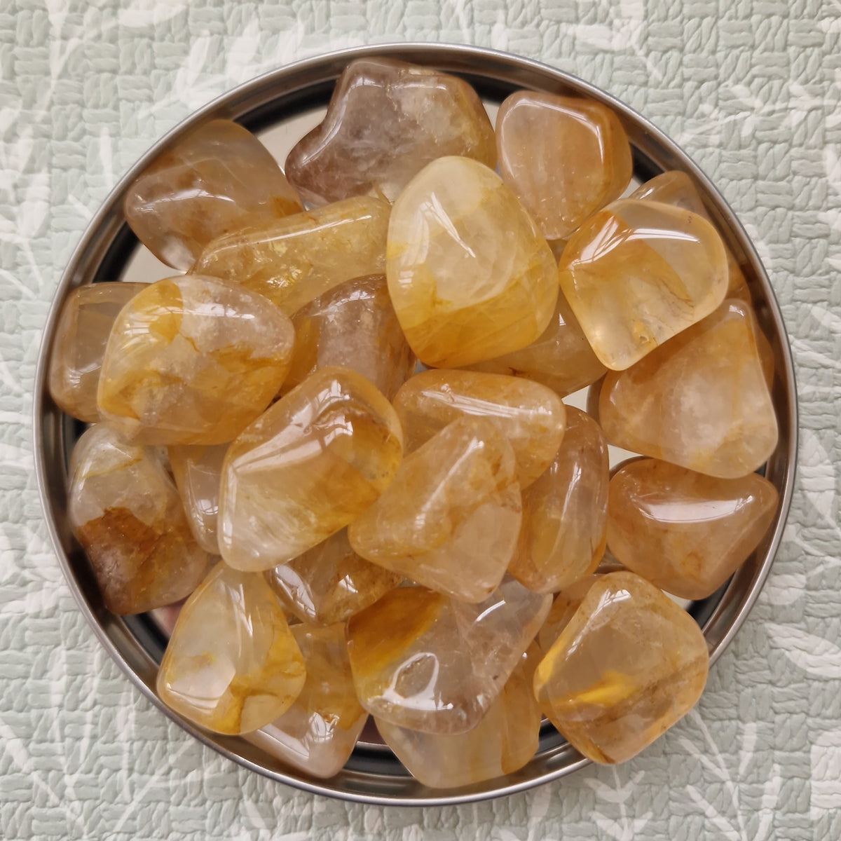 A top-down view of polished tumbled gemstones in warm amber, honey, and light brown tones, arranged inside a round metal dish on a textured light green surface with a white botanical pattern.