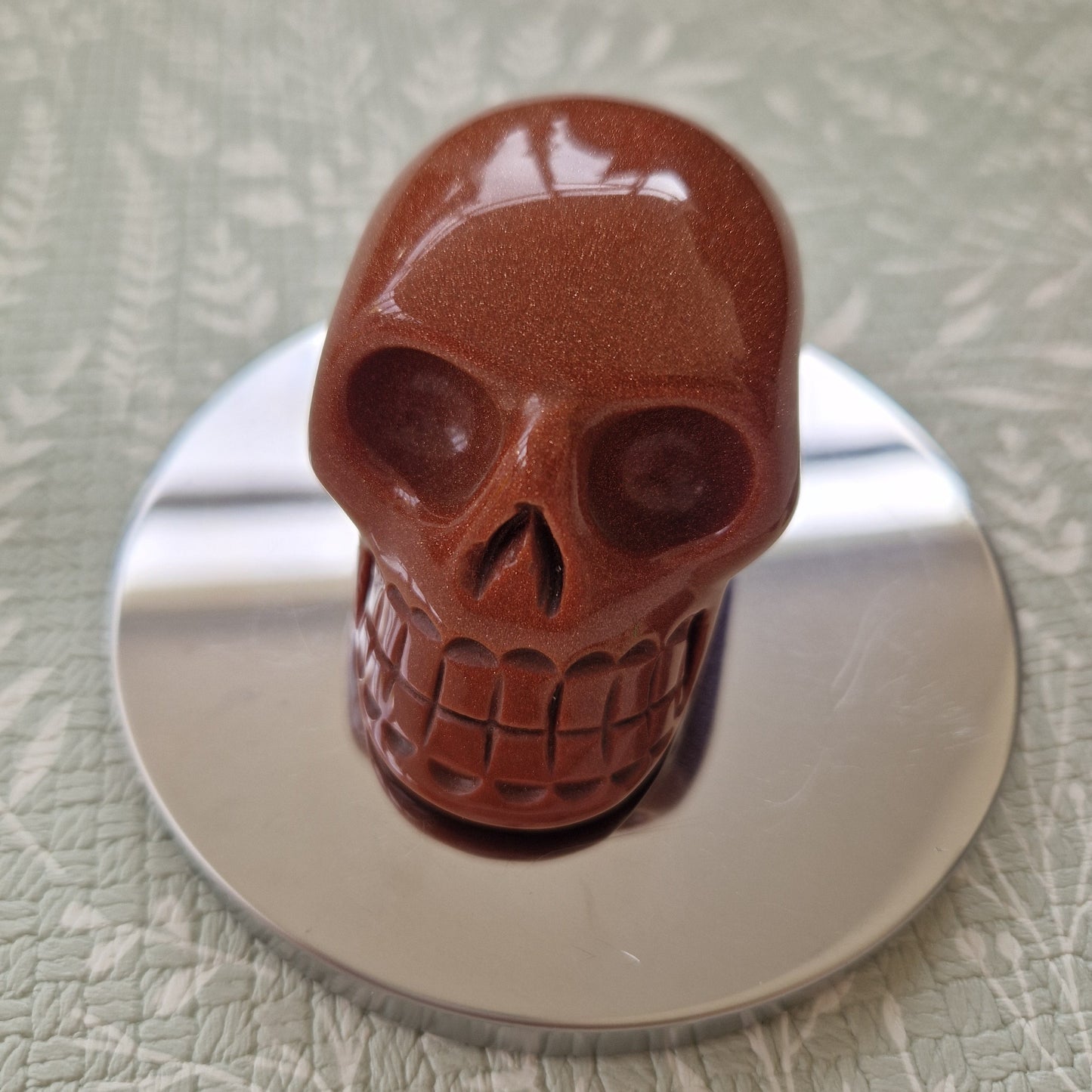 A glossy reddish-brown skull-shaped object displayed on a round silver metal base against a textured pale green background.