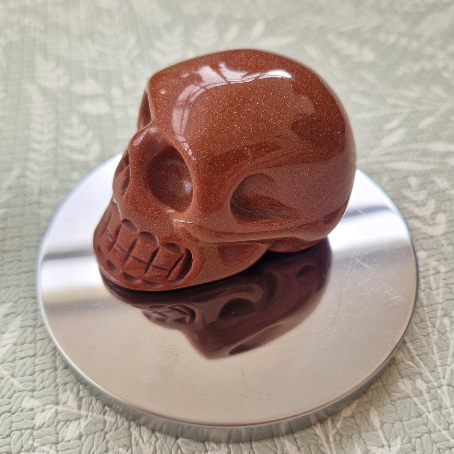 A polished reddish-brown skull sculpture displayed on a round silver reflective coaster over a textured light gray surface.