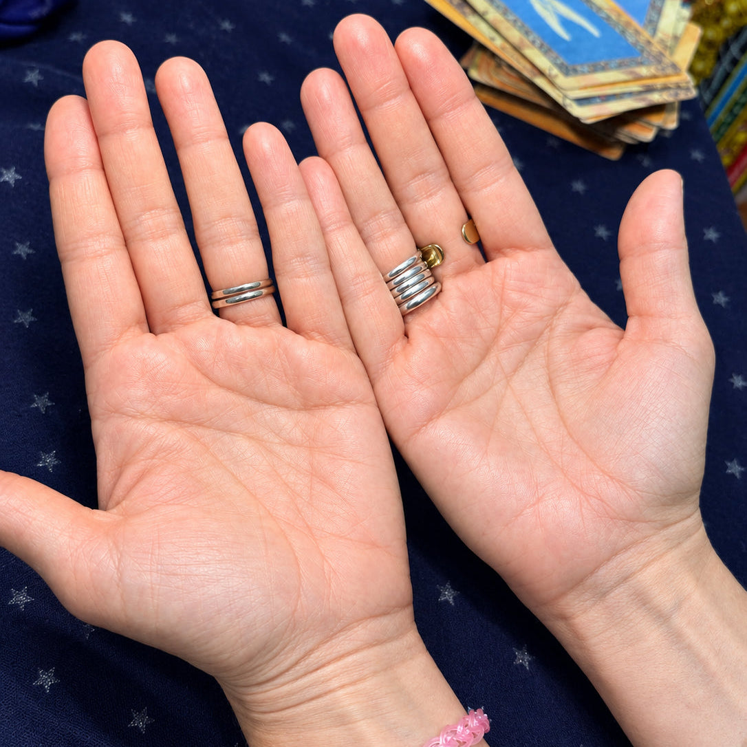 Two open palms showing several silver rings worn on the fingers, with a pink beaded bracelet on one wrist and a dark beaded bracelet on the other, set against a dark blue starry fabric background with tarot cards in the upper right.