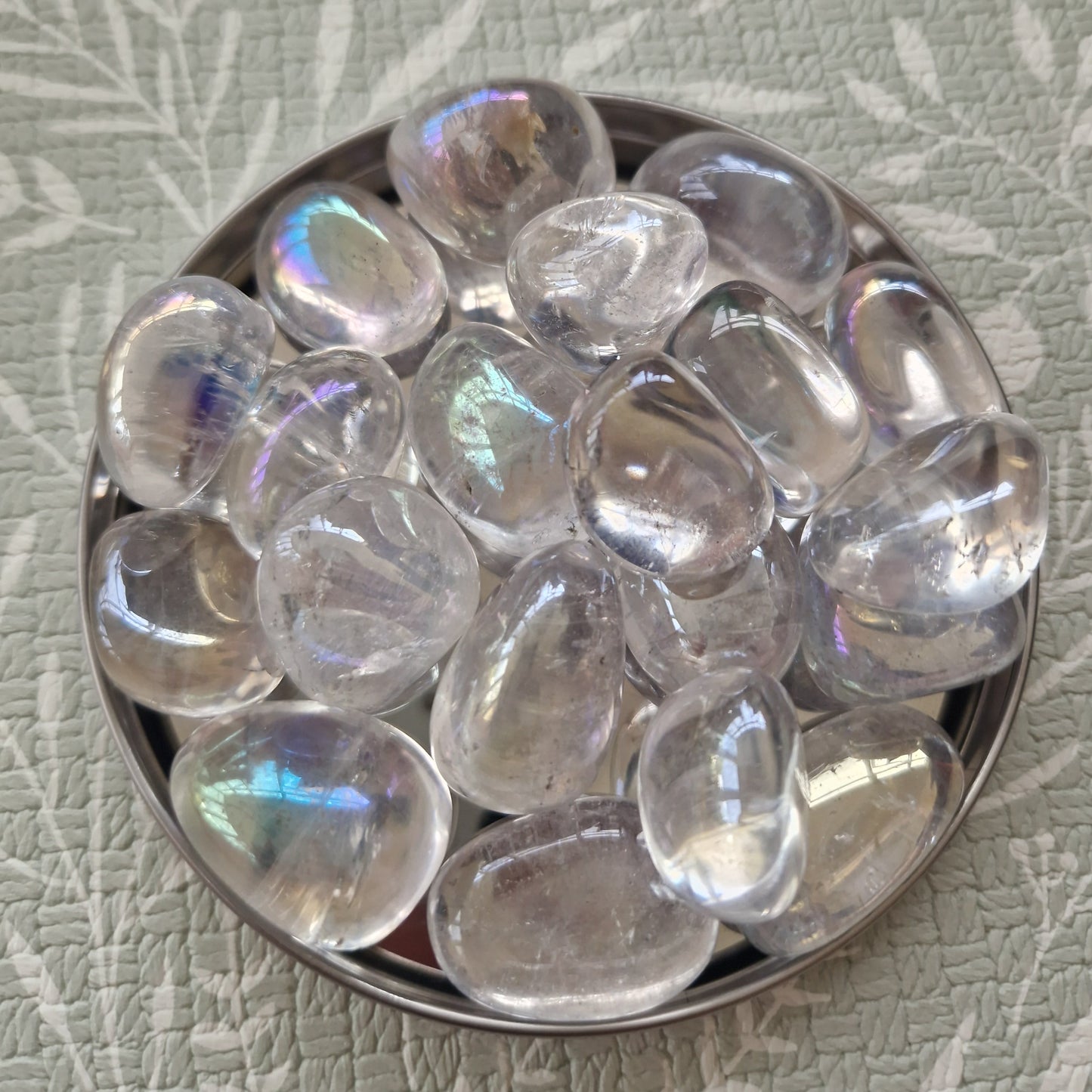 A pile of smooth, polished iridescent glass pebbles in a round metal dish, set against a textured sage green background with a leaf pattern.