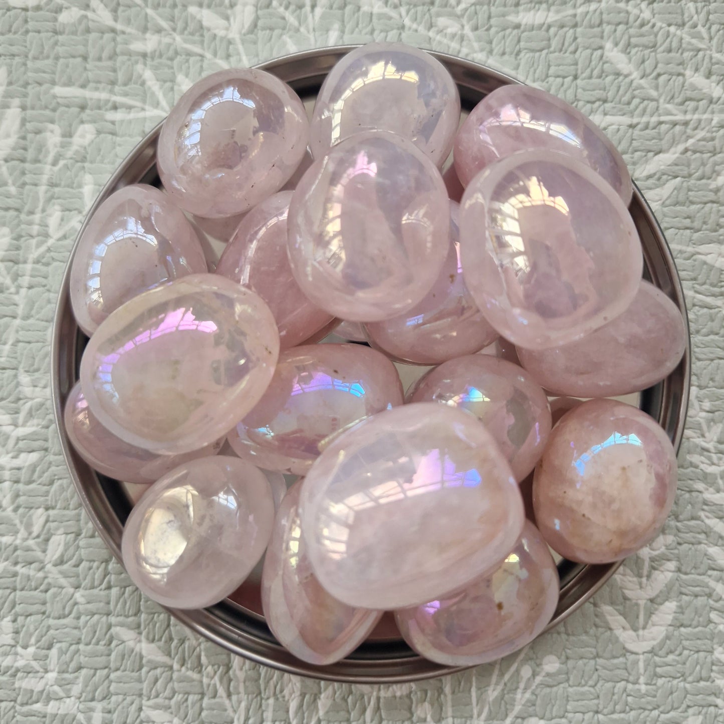 A collection of smooth, polished pink tumbled stones with an iridescent sheen, arranged in a small dark metal bowl on a textured light green background with a white leaf pattern.