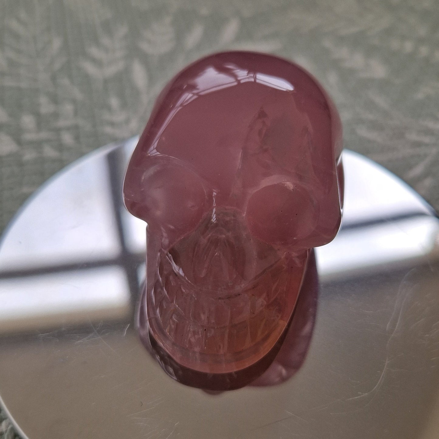 A small translucent pink skull-shaped object displayed upright on a round reflective silver stand, set on a textured light green surface.