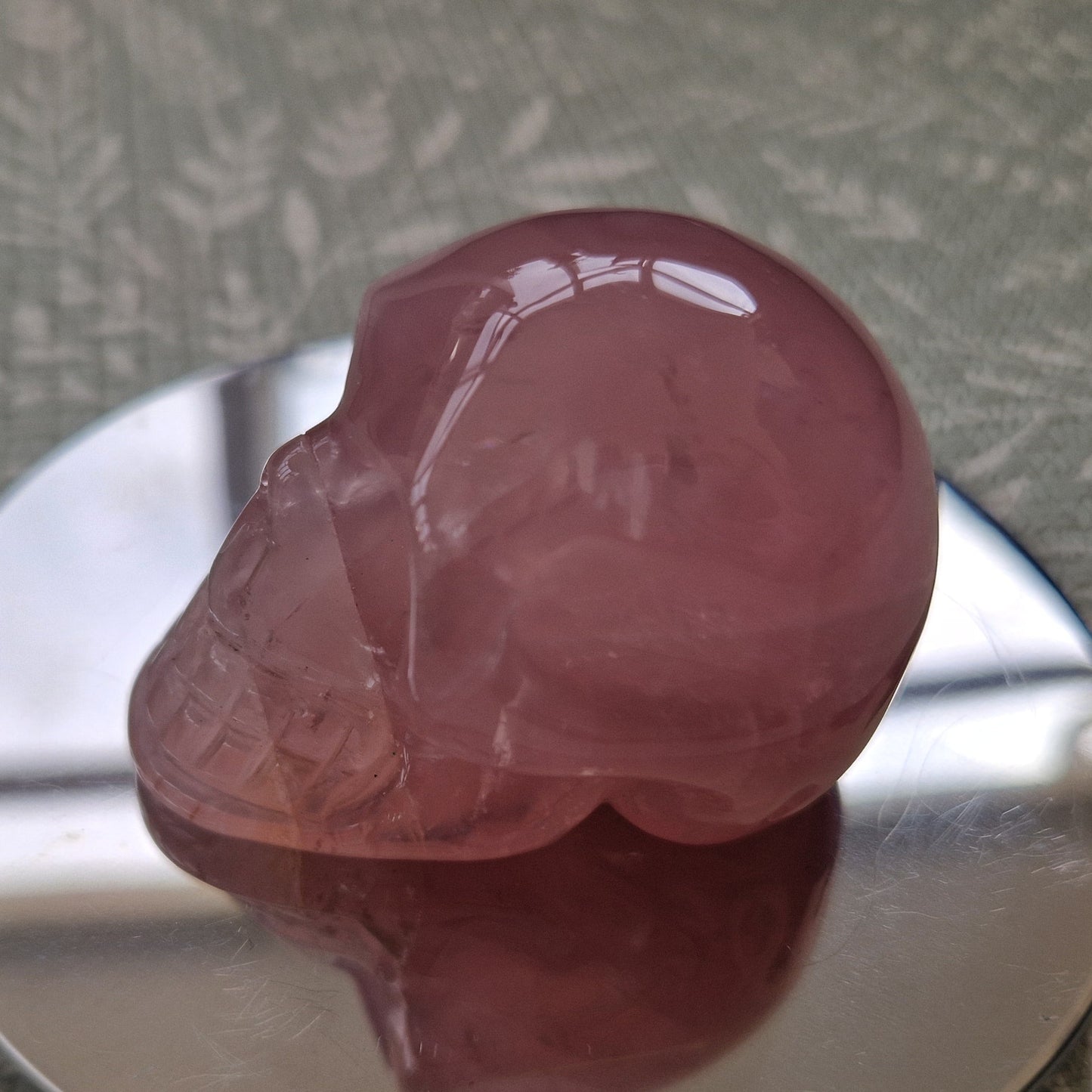 A polished pink crystal skull sculpture displayed on a reflective silver circular stand over a textured green and white patterned fabric surface.