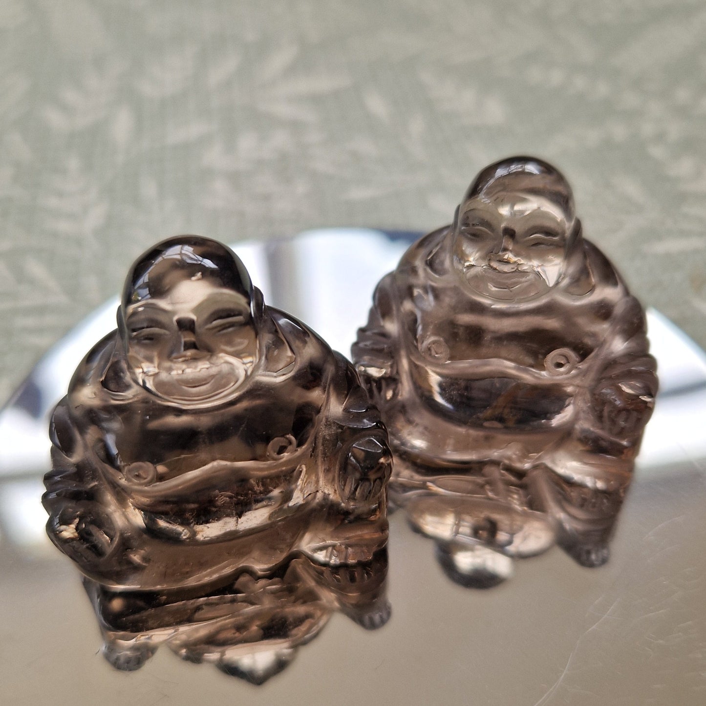 Two small translucent smoky glass Laughing Buddha figurines displayed on a round reflective silver tray over a textured light green surface.