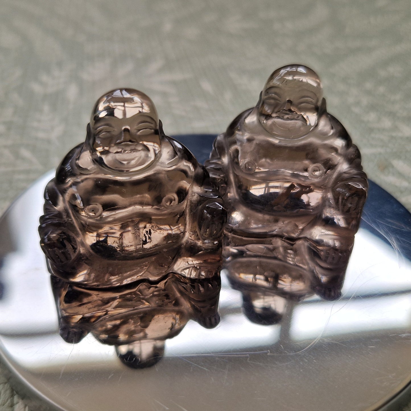 Two small transparent smoky crystal Laughing Buddha figurines sitting side by side on a round reflective silver mirror, placed on a textured sage green surface.